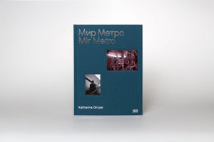 A front view of the photography book Mir Metro by Katharina Gruzei. The square, dark teal, cloth-bound cover features the title Мир Метро and Mir Metro in silver foil. Below the title are two inset photographs: one in black and white of a person on an escalator, and one in color of a reflective, red marbled interior. The book is standing on a white surface.