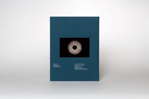 A front view of a hardcover book with a dark teal, textured cloth cover, standing against a plain light grey background. In the center of the cover is a rectangular photograph of a circular, modern light fixture with glowing spokes radiating from a dark center. Below the image, white text lists contributors, including Boris Groys and Abigail Solomon-Godeau.