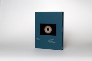 A dark teal hardcover book stands at an angle on a white surface. The front cover features an inset square image of a circular, illuminated object with radiating white lines against a black background, resembling a stylized eye. Silver text is printed on the front cover and the spine.
