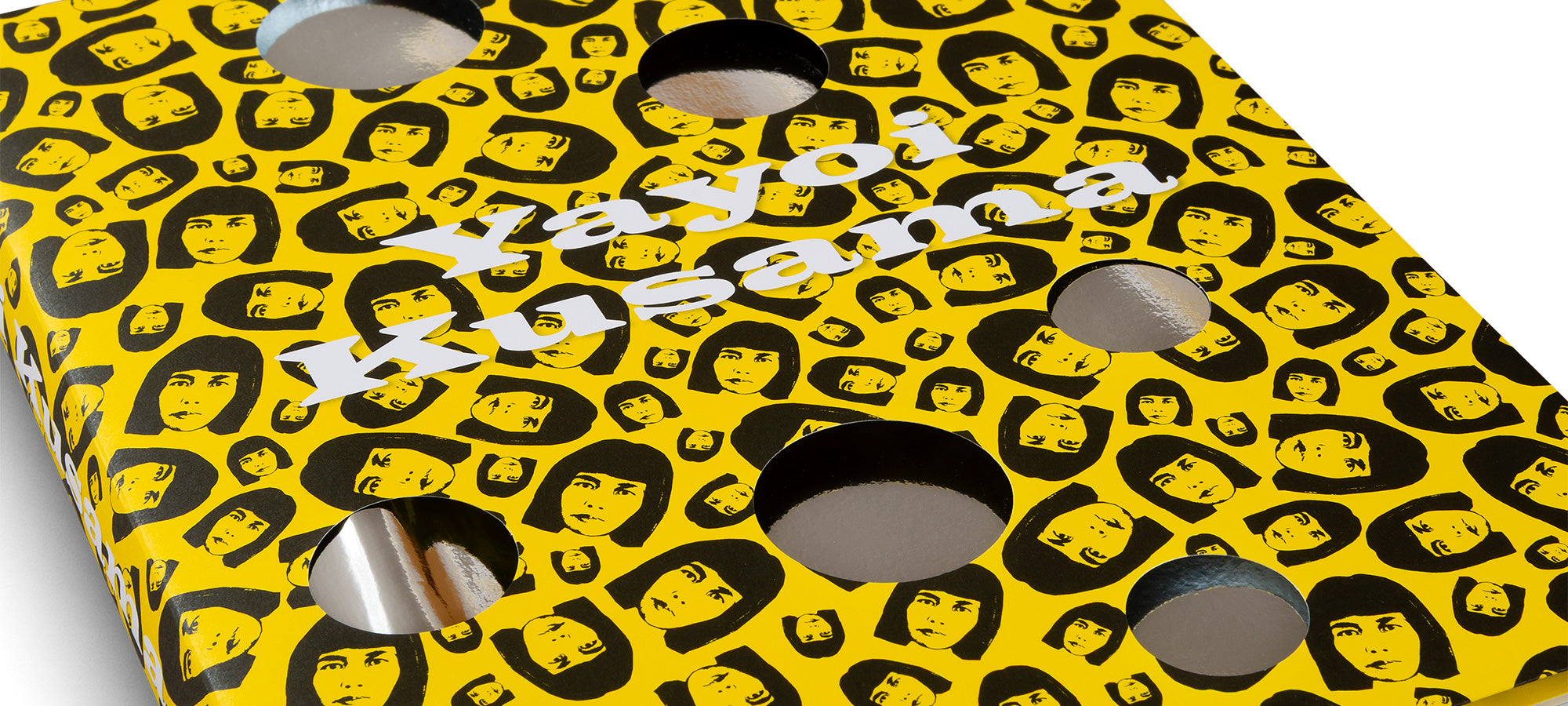 A close-up, angled shot of a bright yellow box covered in a repeating black-and-yellow pattern of a woman's face. The name Yayoi Kusama is written in white across the surface, which is perforated with large, circular cutouts that reveal a shiny silver interior.