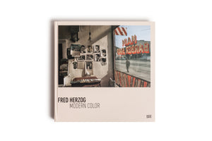 A top-down view of the photography book Fred Herzog Modern Color. The cover features a color photograph taken from inside a vintage barbershop, looking out onto a street. The window reflects the shop's red sign in reverse, reading MAIN BARBER SHOP, and a person walking on the sidewalk. Inside, a wall is covered with old photos and calendars.