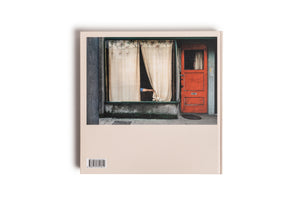 The back of a book with a cream-colored cover, viewed from above against a white background. The cover features a photograph of a building's facade with a weathered, bright red door next to a large window with parted beige curtains. A person's arm in a blue sleeve is visible through the opening in the curtains. A barcode is located in the bottom left corner.