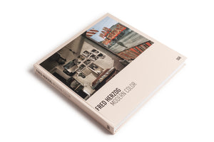A square hardcover book titled Fred Herzog Modern Color, with a pale pink cover, is displayed at an angle on a white background. The cover art is a color photograph showing a vintage interior with old photos on a wall, overlaid with a reflection of a city street and a large red BARBER sign.