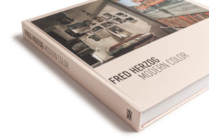 An angled shot of the hardcover book Fred Herzog Modern Color, resting on a white surface. The book has a pale beige cover featuring a photograph of a cluttered desk or wall decorated with vintage papers and old black-and-white portraits.