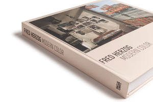 An angled view of the hardcover book Fred Herzog Modern Color on a white background. The book has a pale pink cover with the title in black, modern font. The cover image features a photograph of an interior with vintage photos and papers on a wall, contrasted with a colorful street scene.