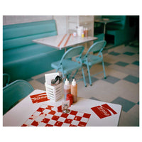 A film-style photo of a retro diner interior. In the foreground, a white table is set with a red and white checkered placemat, Coca-Cola napkins, and a caddy with cutlery and condiments. In the background, a teal vinyl booth, light blue chairs, and another table sit on a gray and blue checkered floor.