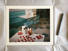 A matted photographic print of a retro diner scene is laid out on white tissue paper next to a gray pen. The photograph shows a table in the foreground with Coca-Cola logos and condiments, with a turquoise booth and chairs on a checkered floor in the background. The print is numbered, titled Table 21, and signed.