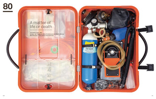 A top-down view of an open, orange hard-shell emergency medical kit, identified as the Ambu case from 1981. The left side has clear compartments for supplies, while the right side is densely packed with resuscitation equipment, including a blue oxygen tank, a manual resuscitator bag, gauges, and tubing.