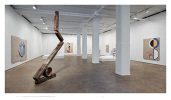 A wide-angle view of an art gallery with white walls, a concrete floor, and white columns. In the foreground, a large sculpture made of weathered wooden beams is suspended in a crooked line. The background features several abstract paintings with colorful geometric circles and other sculptures on white plinths.