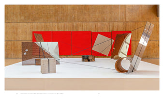 An installation view of an abstract sculpture in a museum. On a white platform, various geometric objects made of wood, glass, and stone are arranged. The composition is set against a wood-paneled wall with a row of bright red panels. Taut black cords are stretched between the objects, creating a web of intersecting lines.