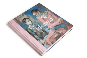 A hardcover book titled Picasso, The Blue and Rose Periods, is shown at an angle on a white background. The cover features a Picasso painting of two solemn figures in costumes against a blue background, and the book has a light pink cloth spine.