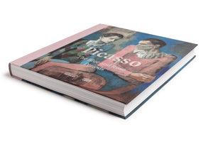 A close-up, angled shot of a hardcover book titled Picasso: The Blue and Rose Periods, set against a white background. The cover features a Picasso painting of two harlequins, one in pink and one in a multi-colored diamond pattern, against a blue backdrop. The book has a pale pink spine.