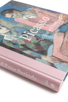An angled view of a hardcover book titled Picasso: The Blue and Rose Periods. The cover features a Picasso painting of two somber figures in blue and pink hues. The book has a textured, pale pink cloth spine with silver lettering.