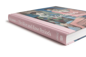 An angled, close-up view of a hardcover book titled Picasso – The Blue and Rose Periods. The book has a light pink, cloth-textured spine with the title in white serif font. The front cover features a painting by Picasso of two figures against a blue background. The book rests on a white surface.