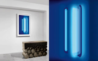 A split image of an art installation. On the left, a framed artwork depicting glowing blue vertical light bars hangs on a white gallery wall above a modern console table made of clustered dark metal pipes. The right side shows a close-up of the artwork, emphasizing the soft, ethereal quality of the blue light against a dark background.
