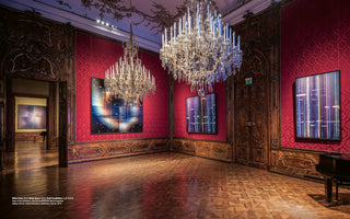 An ornate, baroque-style room with red damask walls, dark wood paneling, and a polished parquet floor, serving as an art gallery. Two large crystal chandeliers hang from the ceiling, illuminating several pieces of modern, luminous abstract art with geometric and digital patterns that contrast with the classical decor. An open doorway reveals an adjacent gallery space.