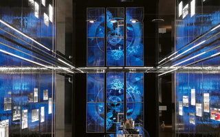 A symmetrical view of a high-tech, modern interior with a central black wall featuring a two-tiered grid of glowing blue digital screens that display abstract watch-like gears and mechanisms. The central display is flanked by dark, reflective, textured walls that shimmer with blue light, all illuminated by bright horizontal LED light strips.