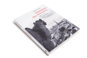 A white hardcover book, Städtebau des Erinnerns by Wolfgang Pehnt, lies on a white surface. The cover features a black and white photo of a stone gargoyle looking out over a cityscape with the Eiffel Tower. The title is printed in red.