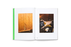A top-down shot of an open photobook with a grey cover and a vibrant green spine. On the left page is a photo of a damaged wood panel wall. On the right page is a photo of a discarded, deflated inflatable doll lying across a trash can and a pile of oranges on a dark city street.