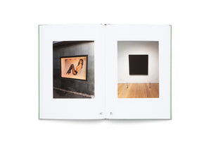 An open book on a white surface, displaying a two-page spread. The left page has a photograph of a lit advertisement of a woman in lingerie on a dark wall. The right page has a photograph of a solid black square painting hanging on a white gallery wall with a wooden floor.