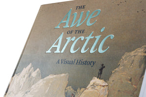 A close-up, angled photograph of the cover of the book The Awe of the Arctic: A Visual History. The title is embossed in a reflective, light-blue foil against a textured, sandy brown background. The cover features a vintage illustration of a person standing on a rocky cliff overlooking a snowy mountain range.