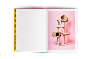 An open book with a colorful cover lies flat on a white surface. The left page is blank, while the right page features a photograph of a calico cat on a multi-level wicker cat tree against a solid pink background.