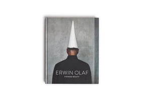 A top-down view of the art book Erwin Olaf: Strange Beauty, resting on a white surface. The cover features a photograph of a person seen from the back, wearing a black shirt and a white conical hat, standing in front of a textured gray wall.