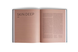 An overhead shot of an open hardcover book on a white surface. The pages are a warm, light brown color. The left page is a chapter title, reading SKIN DEEP in a large, black sans-serif font, with 2015 printed underneath. The right page is filled with two columns of black text.