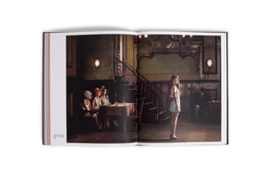 A studio shot of an open art book on a white surface. The book displays a wide photograph of a dimly lit, vintage ballroom. On the left page, three older women in theatrical, burlesque-style costumes sit at a table. On the right page, a young woman in a modern pink top, light skirt, and purple platform heels stands alone, looking back over her shoulder. The scene creates a stark contrast between generations and styles.