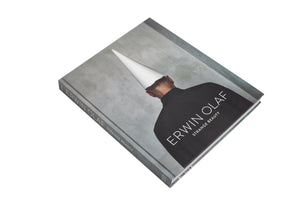 A grey hardcover book titled Erwin Olaf: Strange Beauty, laid diagonally on a white background. The cover features a photograph showing the back of a person wearing a black shirt and a white conical hat against a grey, textured wall.