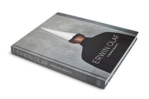A gray hardcover book titled Erwin Olaf: Strange Beauty, resting on a white surface. The cover art displays the back of a person in a black jacket, whose head is obscured by a white pointed cone, set against a concrete-like gray background.