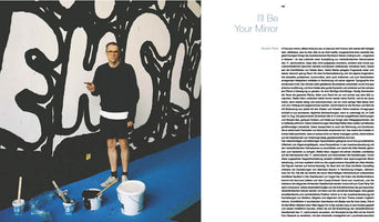 A magazine spread shows a photo on the left of an artist with glasses and black clothing standing in front of a large wall covered in a bold, black-and-white abstract mural. On the blue floor are several buckets of paint and brushes. The page on the right has the title I'll Be Your Mirror above a block of German text.