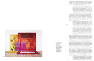 A page from an art book showing an installation of several artworks on a wooden floor. A large red abstract color field painting and a print of Van Gogh's Sunflowers stand against a white wall. In front of them are two smaller, vibrant pink and purple abstract pieces. The right side of the page has a column of German text.
