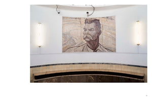 A large mosaic portrait of a man with a prominent mustache is centered on a white, curved wall in a public station. The artwork is flanked by two vertical, glowing light fixtures. Below the portrait is a tiled archway, and two security cameras are mounted on the wall above the mosaic.