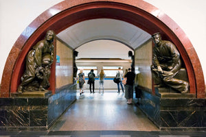 An interior view of a grand metro station looking through a large, red and black marble archway. The arch is flanked by two large bronze statues in niches, one of a person tending to a wounded comrade and the other of a seated sailor. Through the arch, a group of passengers stands on a platform waiting for a subway train.