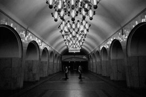 A moody, black and white photograph looking down the length of a grand, vaulted subway station hall. A massive, intricate chandelier composed of glowing rods and spheres hangs centrally, illuminating the arched alcoves that line the polished walls. The vast space is empty except for two lone figures standing in the shadows at the far end.