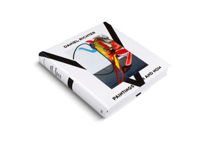 A high-angle studio shot of the art book Daniel Richter: Paintings Then and Now. The thick, white hardcover book is displayed on a white background. The cover features a vibrant, abstract painting with prominent red, yellow, and blue colors, framed by bold black graphic lines.