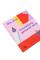 A photograph of a German book titled Wie eine Ausstellung gemacht wird, lying at an angle on a white surface. The pink cover features colorful geometric shapes and whimsical illustrations of a character on a smiling orb and a dog on a rock.