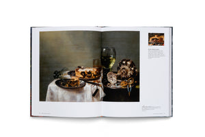 An open art book displaying a two-page spread. The left page is a full-page reproduction of a Dutch still life painting of a lavish table setting with a half-eaten pie, wine, an ornate nautilus cup, and an overturned silver goblet. The right page contains German text and a smaller inset photo.