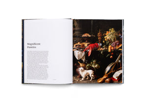 An open art book on a white surface. The left page has text under the heading Magnificent Pantries. The right page shows a dark, opulent still-life painting of a banquet table laden with a large red lobster, pies, fruit, and a golden goblet, with a small white dog and a violin below.