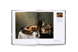 An overhead shot of an open art book. The left page displays a full-page reproduction of Willem Claesz. Heda's still life painting, Breakfast Table with Blackberry Pie. The right page features descriptive text and a smaller detail shot of the painting. The book rests on a flat surface.