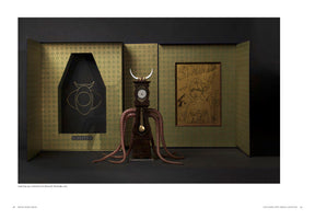 An art installation on a black surface featuring a grandfather clock modified into a surreal monster with white horns and numerous brown, octopus-like tentacles. The creature stands before a three-panel screen with a green and gold pattern. On the left panel is a black recess with a gold bull-like symbol above the word SUMMONING. On the right panel hangs a framed, gold-toned line drawing of a woman.