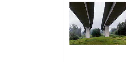 A two-page spread with a blank left page and a photograph on the right. The photo shows a low-angle view from underneath two parallel, curving concrete overpasses supported by large pillars, stretching across a green, forested landscape on a foggy day.
