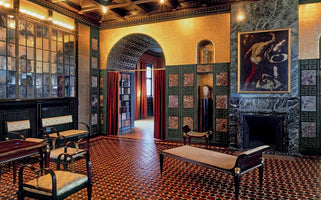 An opulent, dimly lit room with a geometric tile floor and walls decorated with gold mosaics and green marble. On the right is a large marble fireplace with a classical painting above it. In the center, an arched doorway with red curtains reveals a library. On the left, a large, multi-paned mirror hangs above a seating area with dark wood chairs.