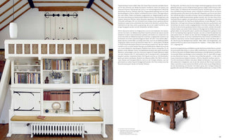 A page from a book showing an Arts and Crafts style room with a high, vaulted ceiling and exposed wooden beams. A large, white, built-in settle with a yellow cushion dominates the left wall, incorporating cabinets and bookshelves filled with books and ceramics. To the right stands a heavy, round, dark wood table on a carved base. The image is on the left page of a spread, with a column of German text on the right.