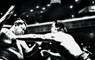 A grainy, high-contrast, black and white action photograph of a sumo wrestling match. A wrestler on the right extends his arm in a powerful thrust, making contact with the face and neck of his opponent on the left. The opponent reels back from the impact, his eyes squinted and one arm raised, capturing the raw intensity of the sport.