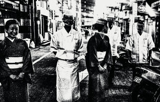 A grainy, high-contrast black and white photograph of five people on a Japanese city street. Two women in kimonos and three men in white uniforms, possibly chefs, stand together. The harsh lighting and deep shadows obscure their faces, giving the scene an eerie, artistic quality.