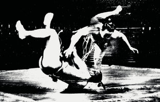 A grainy, high-contrast black and white photograph of two wrestlers captured in a blur of intense motion. One wrestler is upside down in mid-air, being thrown by the other. The image is abstract and chaotic due to the significant motion blur, with the white figures starkly contrasted against the dark background.