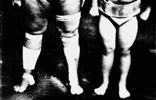 A grainy, high-contrast, black and white photograph of the lower bodies of two figures standing side by side, likely sumo wrestlers. The larger figure on the left has muscular legs heavily wrapped in white bandages around the knee and ankle. The much smaller figure on the right stands with bare legs, wearing a mawashi.