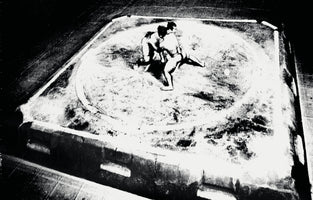 A grainy, high-contrast black and white photograph from a high angle shows two sumo wrestlers mid-match, grappling inside a circular dohyō on a raised square platform.